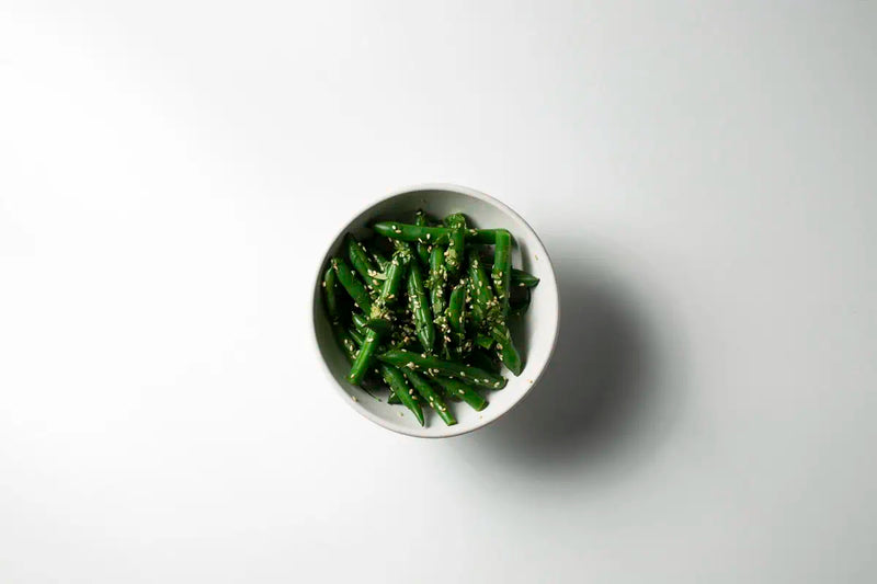 Cooked green beans sprinkled with sesame seeds served in a white ceramic bowl on a clean white background with a soft shadow