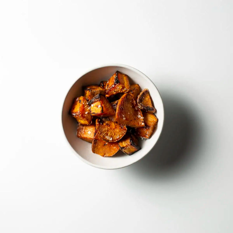 Roasted glazed sweet potato chunks served in a small white bowl on a clean white background, viewed from above