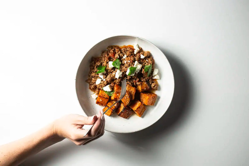 Sweet potato and ground meat bowl topped with white cheese cubes, served in a white ceramic bowl with a fork on a light gray background
