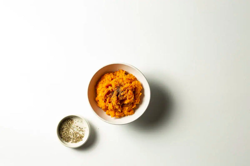 Orange mashed sweet potato or pumpkin with a side of seasoning served in a white bowl on a clean white background