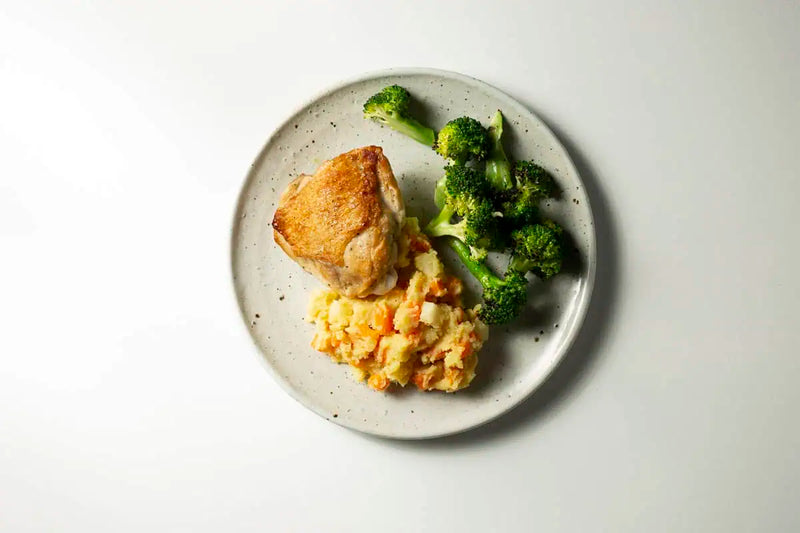 Roasted chicken with broccoli florets and seasoned mashed potatoes served on a speckled ceramic plate against a clean white background