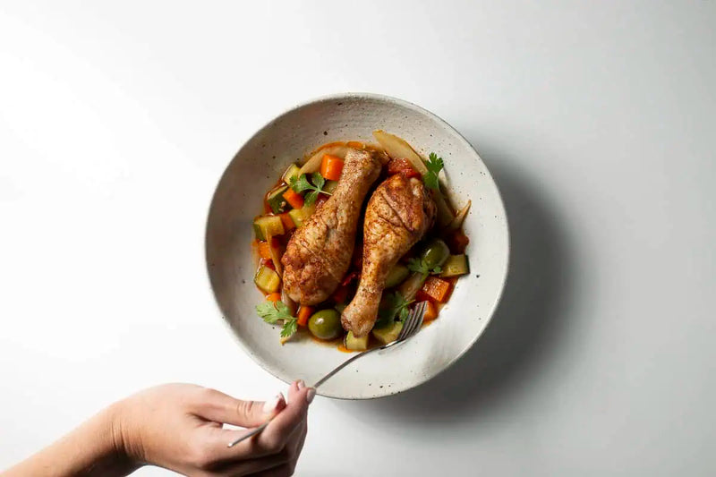 Cooked chicken legs served over a hearty vegetable stew in a ceramic bowl, with a fork on a light gray background